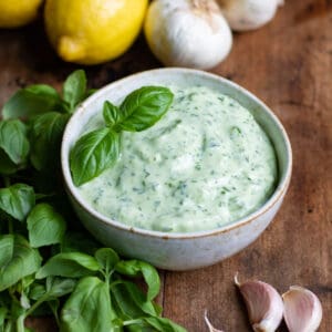 Wooden table with bowl of basil aioli, basil leaves, cloves of garlic and lemons.