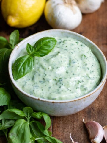 Wooden table with bowl of basil aioli, basil leaves, cloves of garlic and lemons.