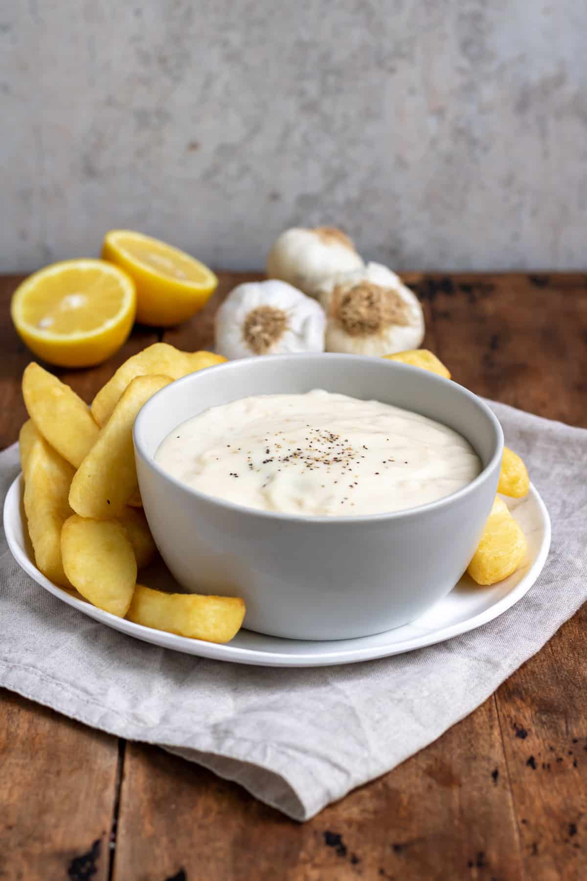 Table with a plate of fries, bowl of horseradish aioli, slices of lemon and cloves of garlic.