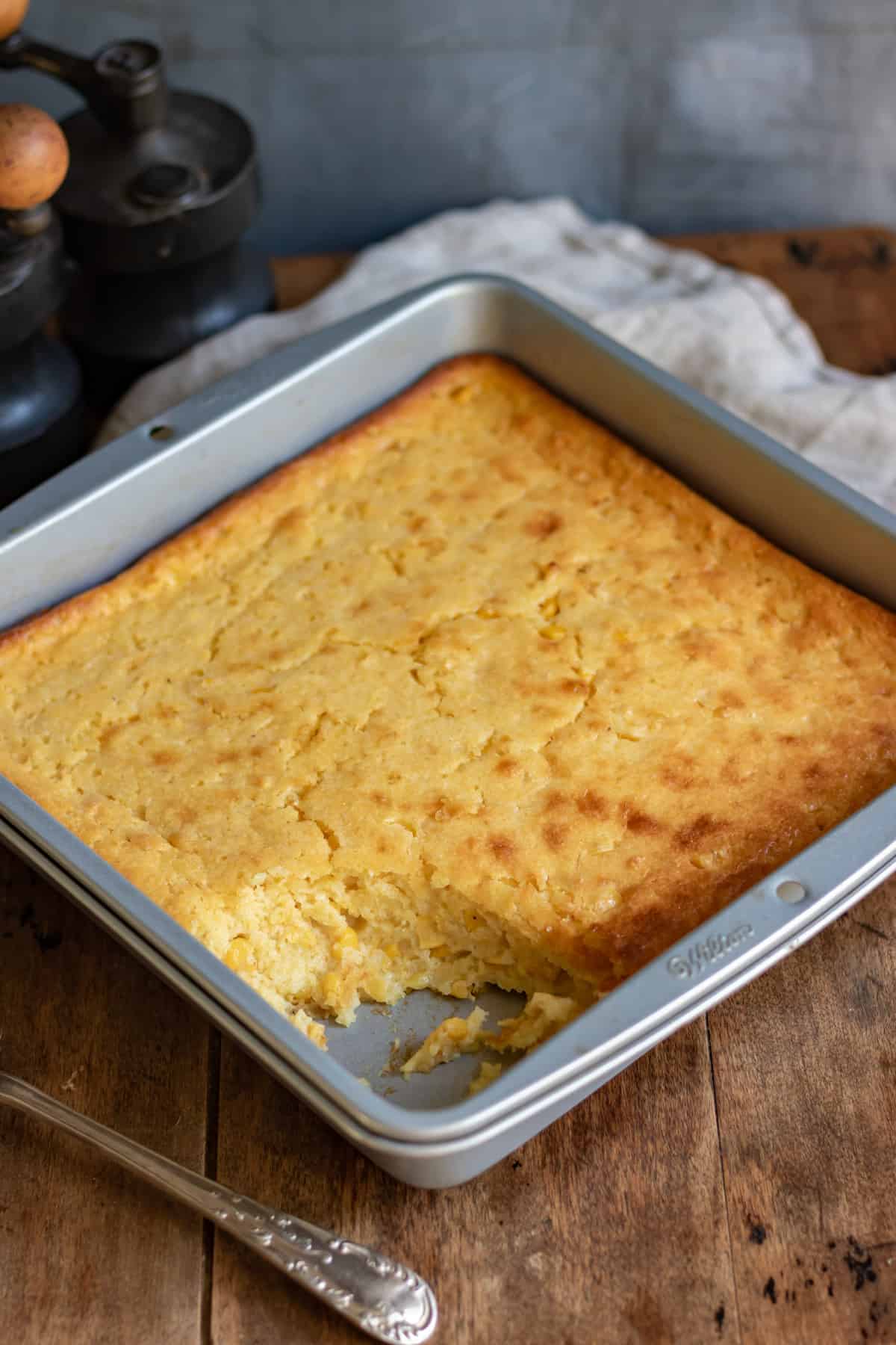Pan of Jiffy corn casserole with a portion removed, on a wooden table.