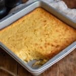 Pan of Jiffy corn casserole with a portion removed, on a wooden table.
