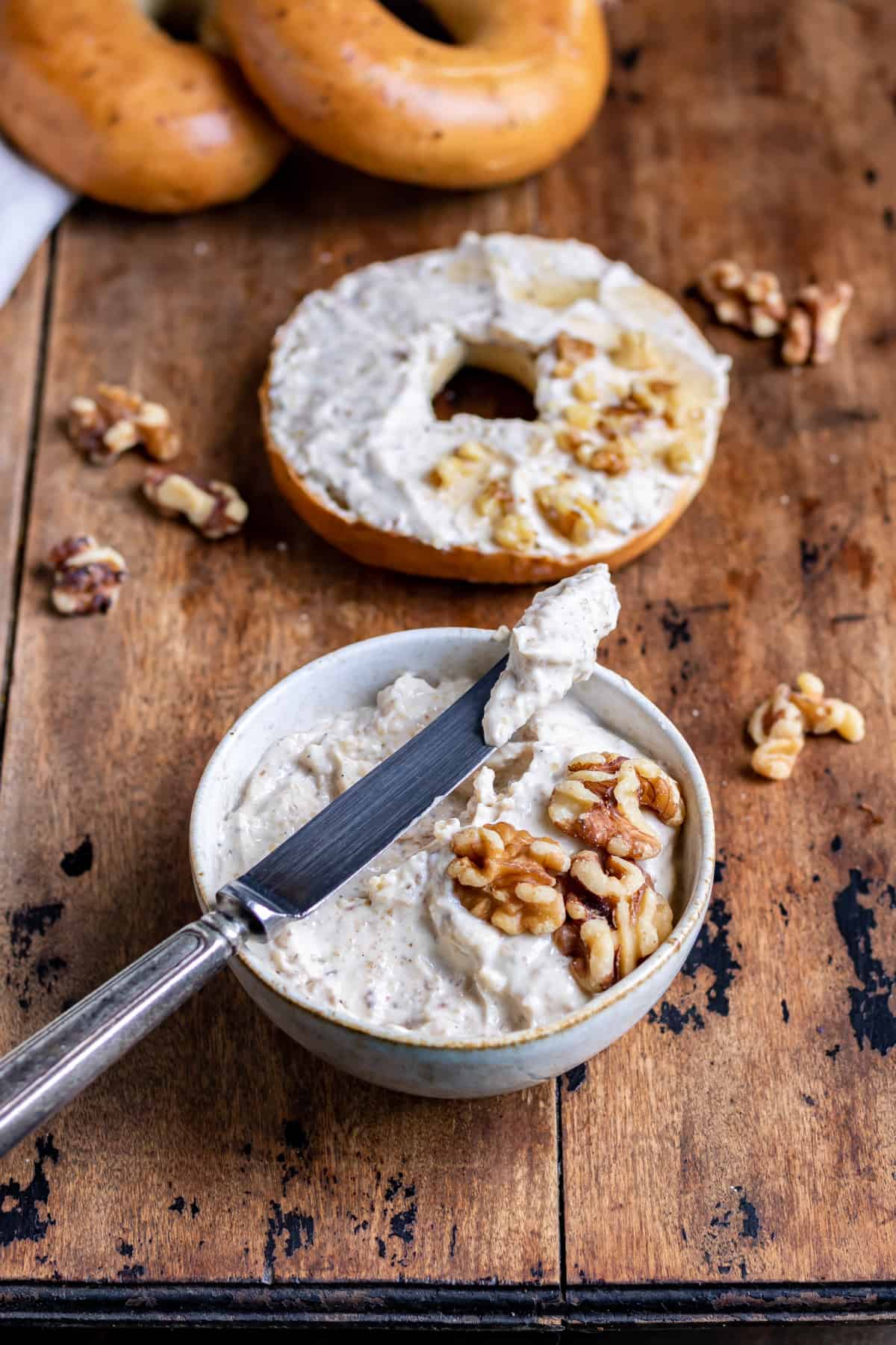 Table with a bowl of honey walnut cream cheese with a knife resting on it and a bagel in the background.