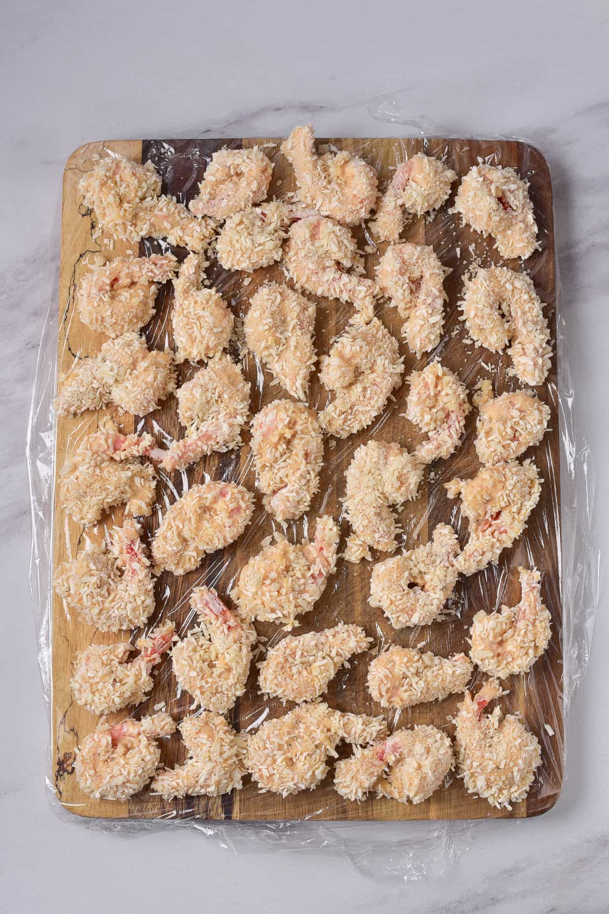 Coated shrimp laid out on a wooden board lined with plastic wrap.