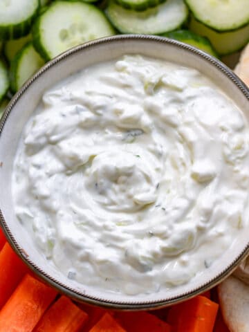 Looking down at a bowl of tzatziki sauce next to carrot sticks, cucumber slices and pita bread.