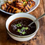 Close up of a bowl of teriyaki sauce with sesame seeds and sliced scallions in it, in front of a plate of chicken and rice.