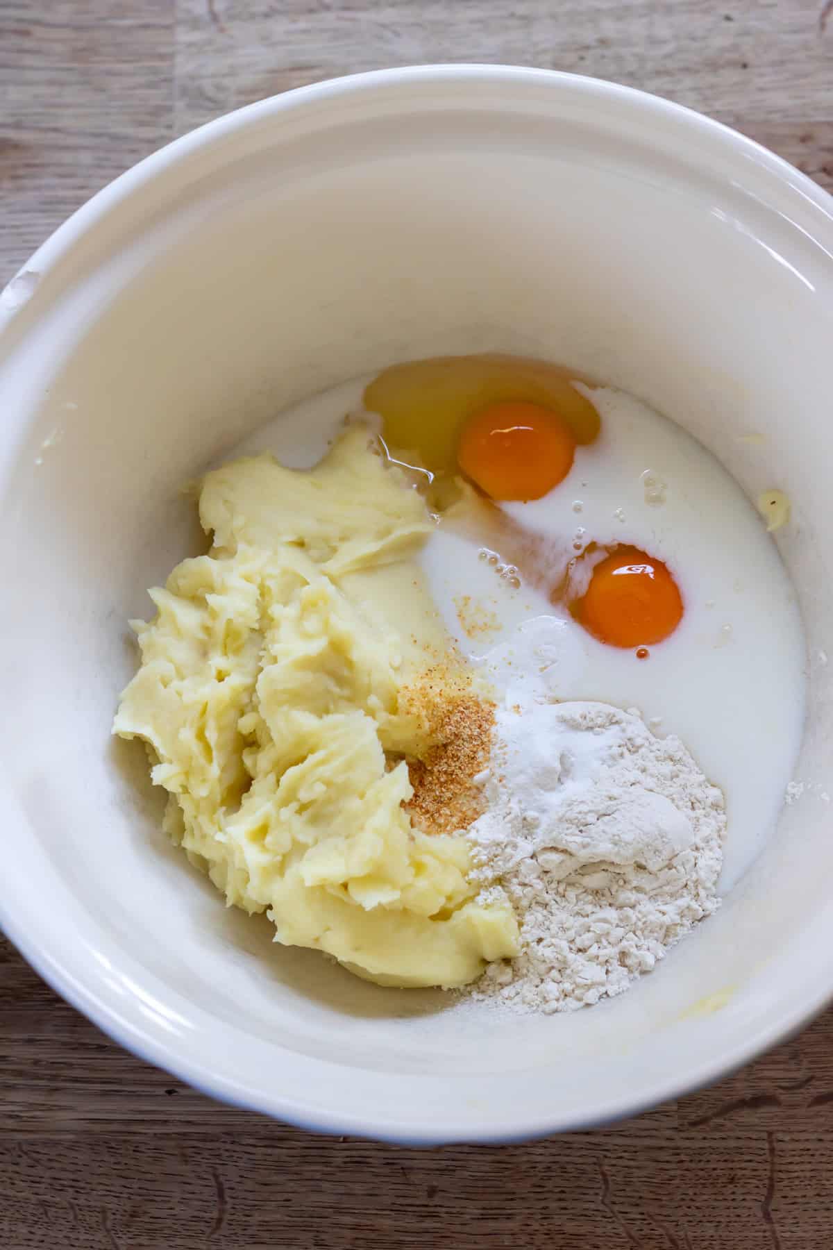 Mashed potato, flour, seasonings, eggs and milk in a mixing bowl.