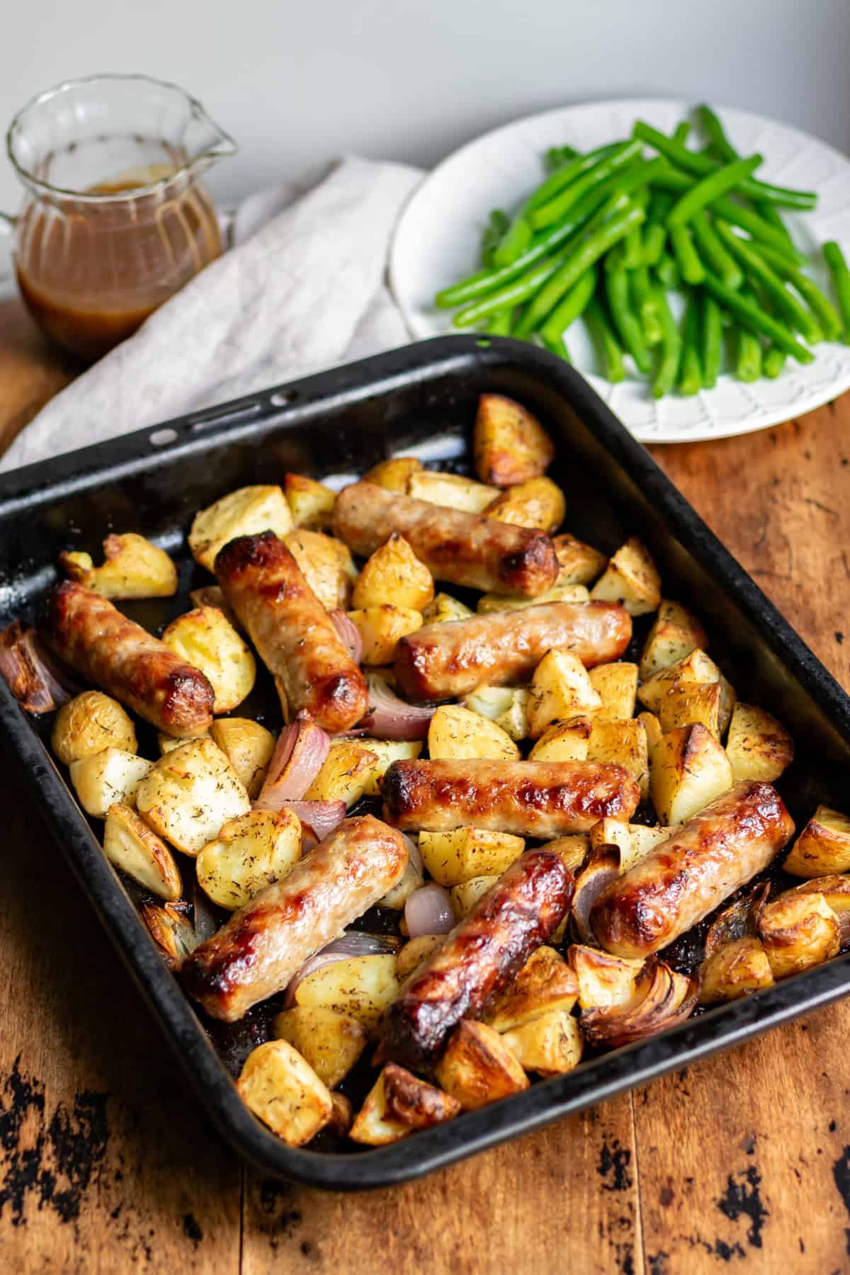 Wooden table with a baking pan of potato sausage bake, next to a jug of gravy and plate of green beans.