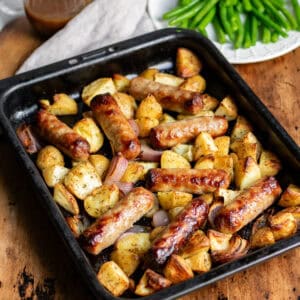 Wooden table with a baking pan of potato sausage bake, next to a jug of gravy and plate of green beans.