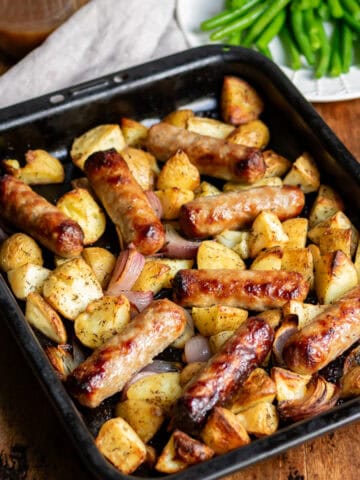 Wooden table with a baking pan of potato sausage bake, next to a jug of gravy and plate of green beans.