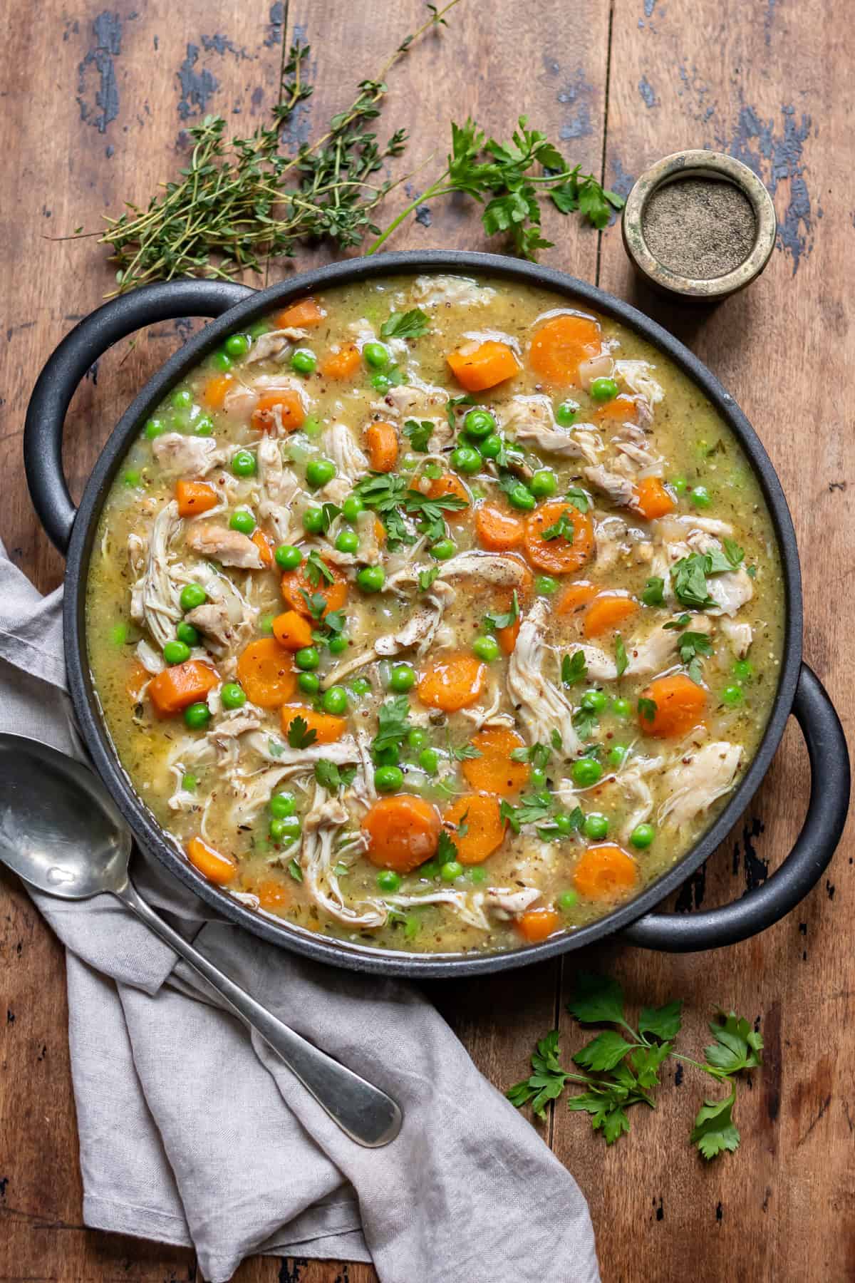 Wooden table with a dish of slow cooker chicken casserole, next to a napkin, pot of pepper and some herbs.