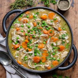 Wooden table with a dish of slow cooker chicken casserole, next to a napkin, pot of pepper and some herbs.