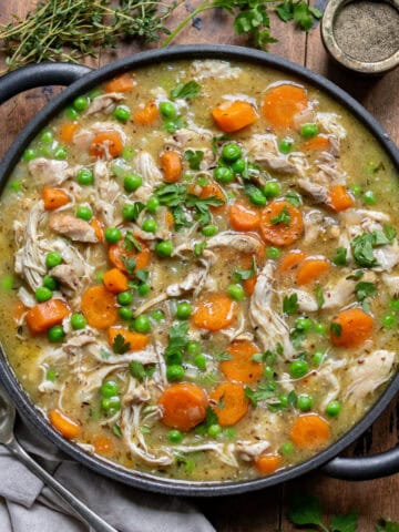 Wooden table with a dish of slow cooker chicken casserole, next to a napkin, pot of pepper and some herbs.