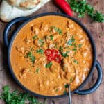 Spoon in a serving dish of slow cooker chicken tikka masala topped with coriander, onion seeds and chopped red chili.