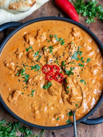 Spoon in a serving dish of slow cooker chicken tikka masala topped with coriander, onion seeds and chopped red chili.