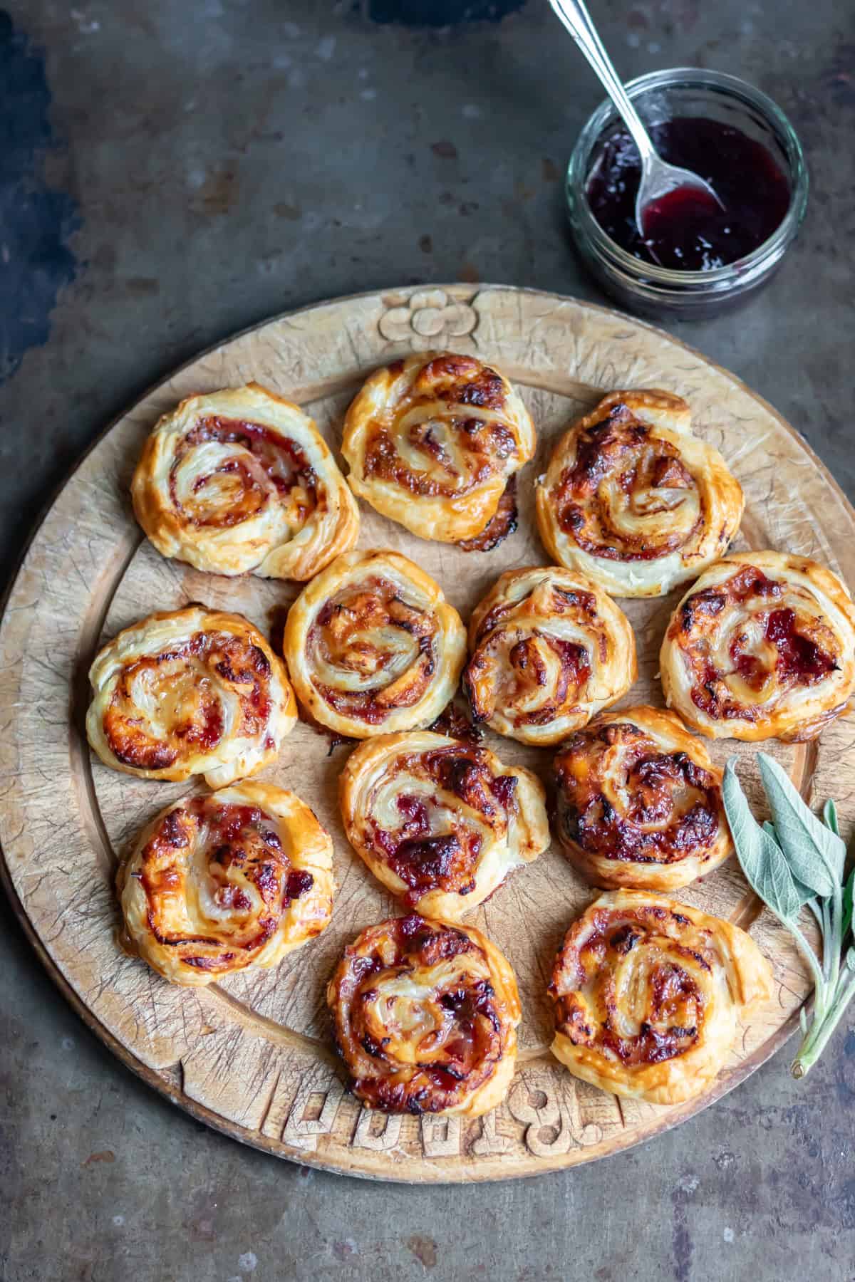 A wooden cutting board of cranberry brie pinwheels next to a piece of sage and a dish of cranberry sauce.