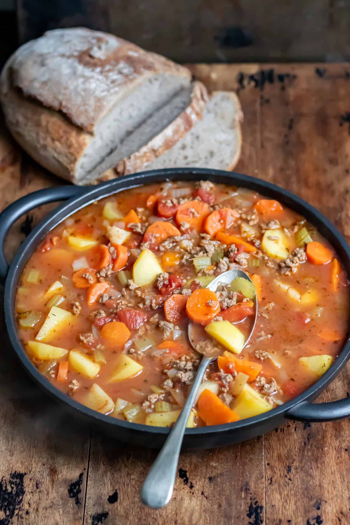 Wooden table with a serving dish of ground beef and potato soup, next to a loaf of bread.