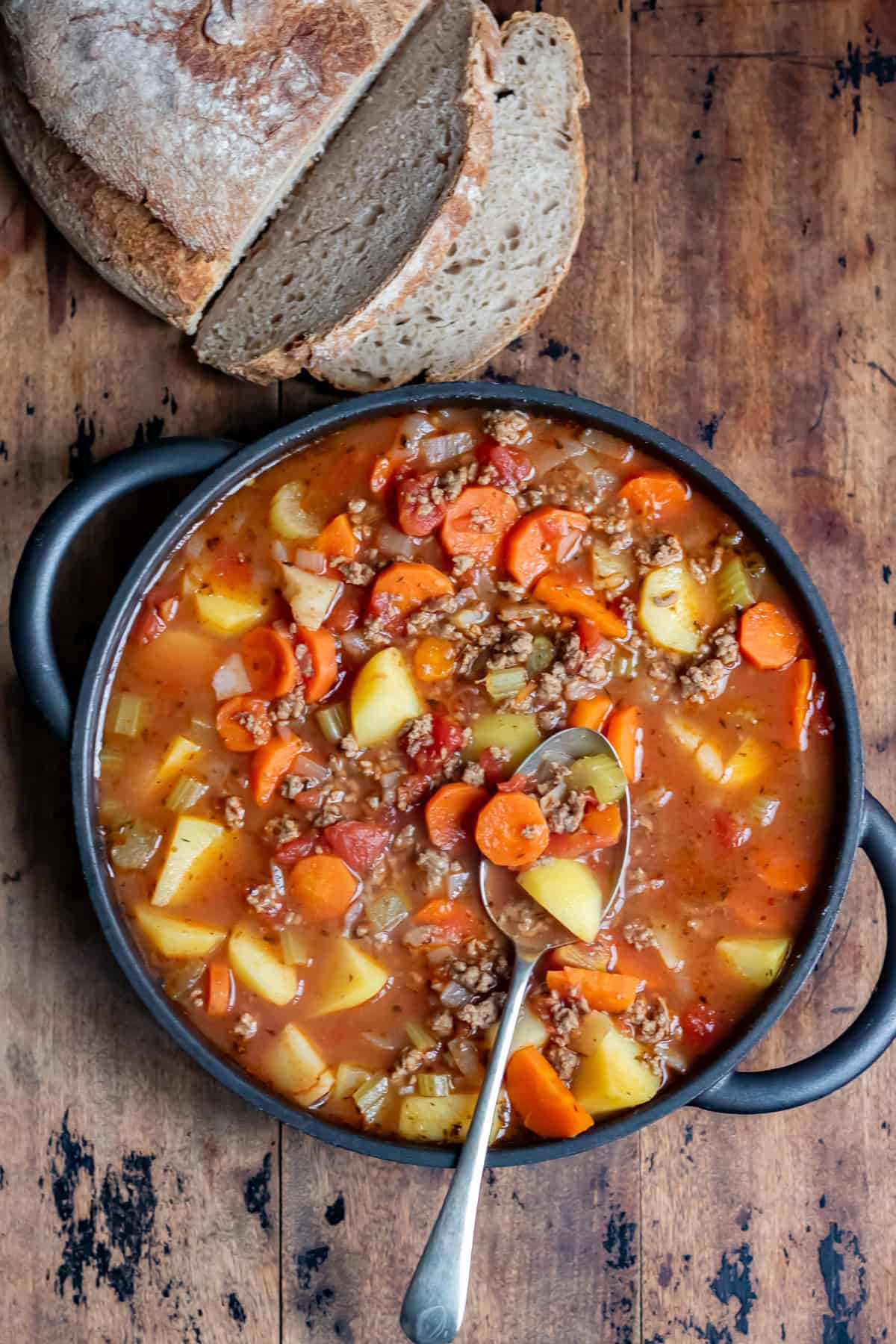 Looking down at a table with a serving dish of ground beef soup next to a sliced loaf of sourdough bread.