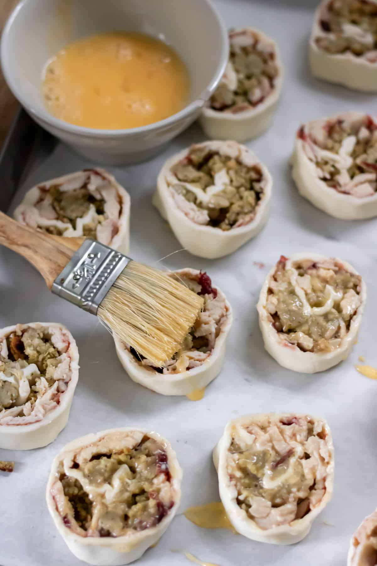 The pinwheels placed onto a baking sheet lined with baking paper, being brushed with beaten egg.