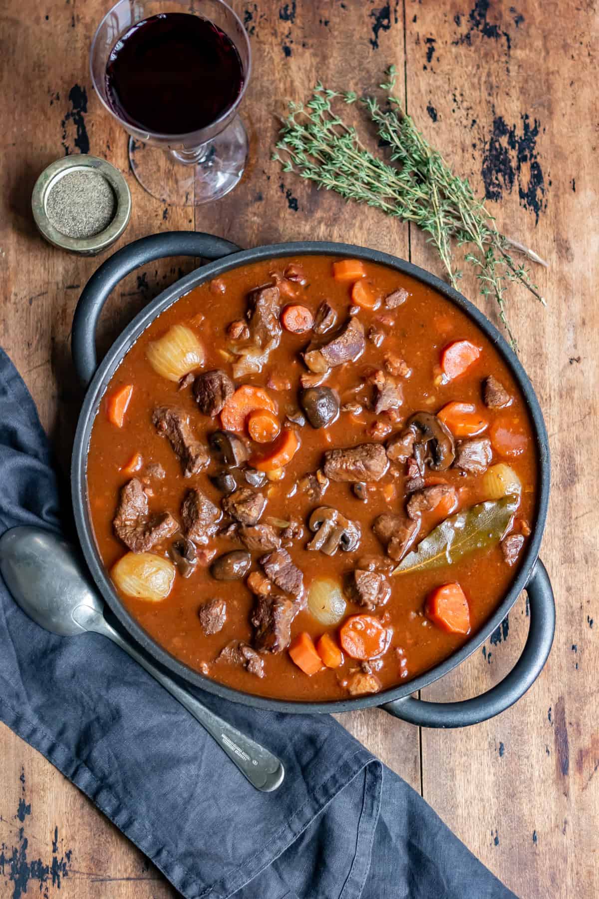 Looking down at a wooden table with a glass of wine, sprigs of thyme and a serving dish of slow cooker beef bourguignon.