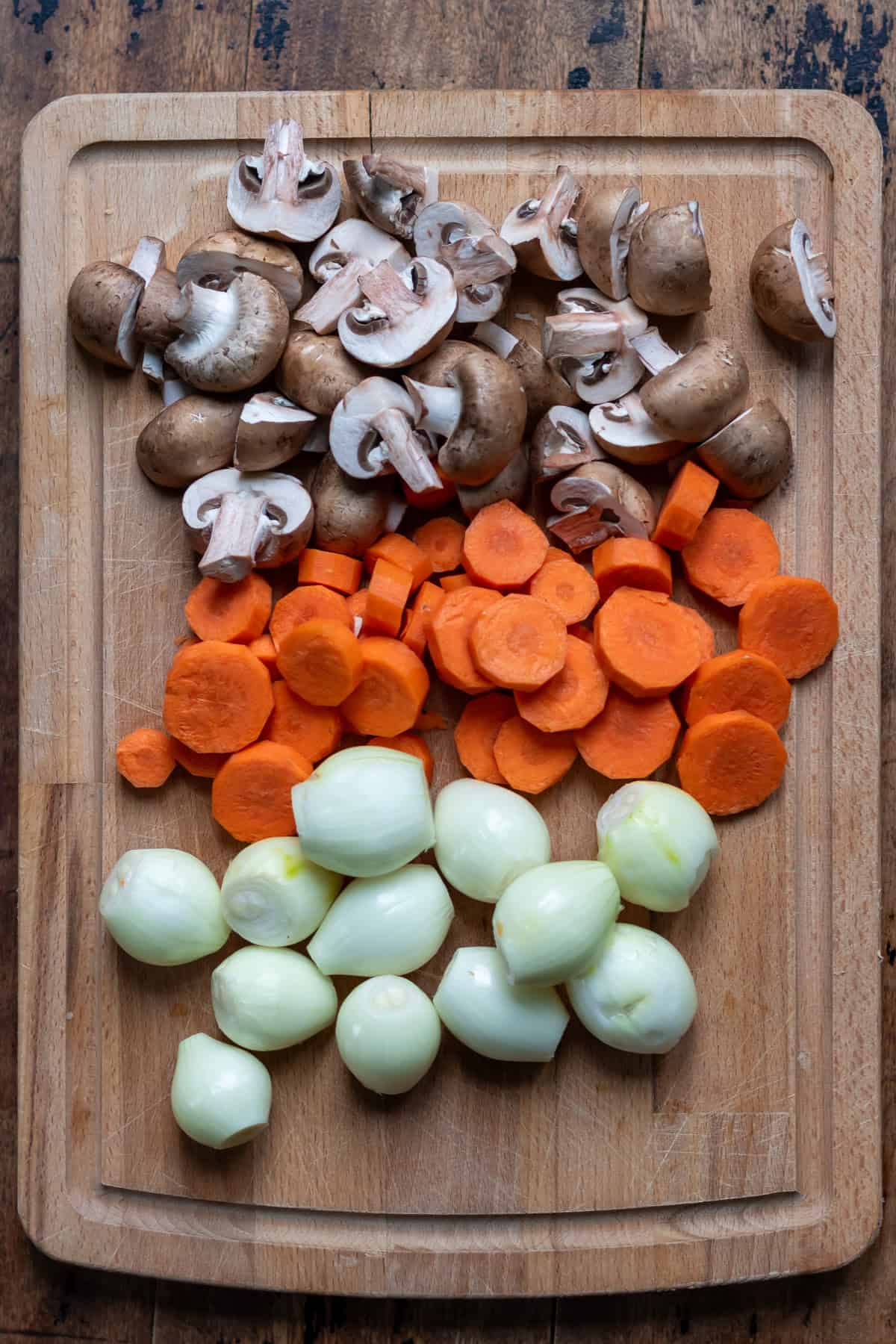 Cutting board with chopped mushrooms, carrots and peeled baby onions.