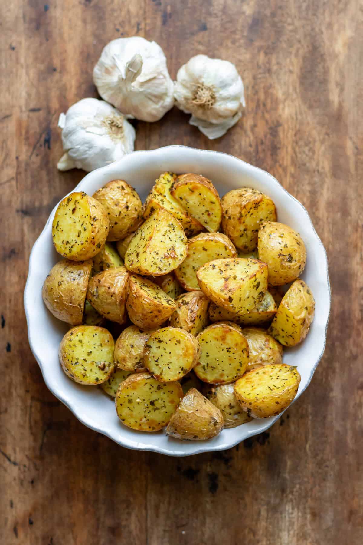 A wooden table with a bowl of air fryer garlic baby potatoes next to a few bulbs of garlic.