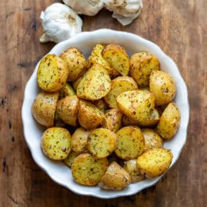 A wooden table with a bowl of air fryer garlic baby potatoes next to a few bulbs of garlic.