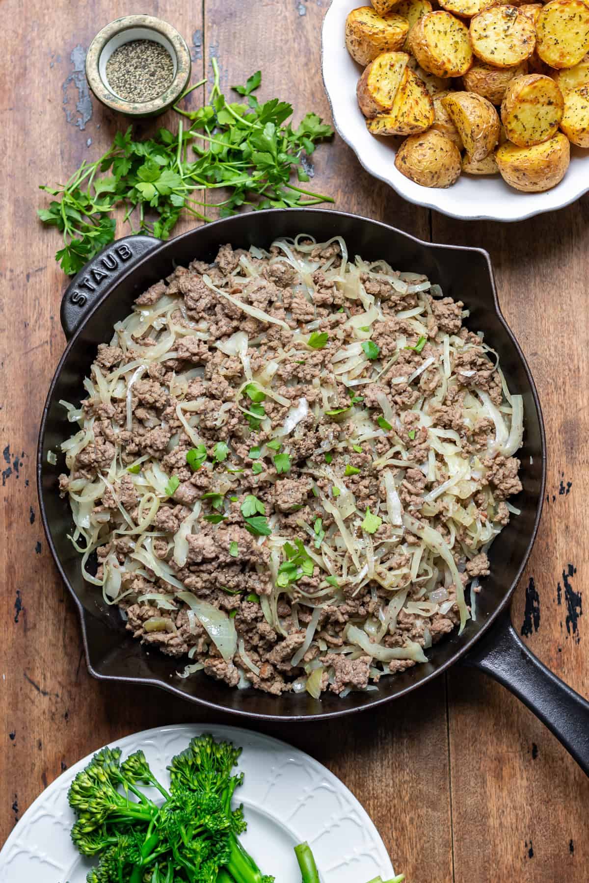 A skillet of ground beef and cabbage on a table, next to dishes of potatoes and broccoli.