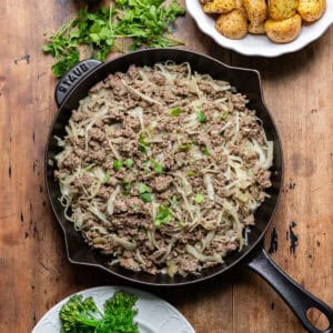 A skillet of ground beef and cabbage on a table, next to dishes of potatoes and broccoli.