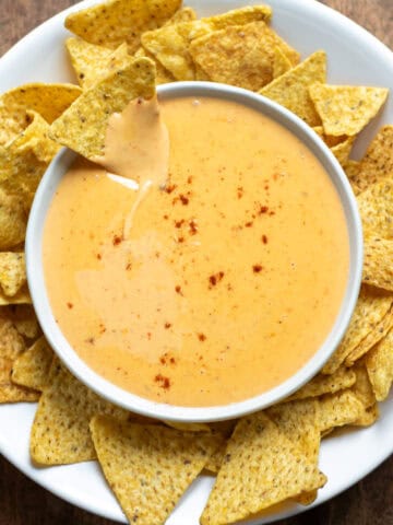 A table with a bowl of nacho cheese sauce, with one tortilla chip in the dip, and surrounded by more chips.