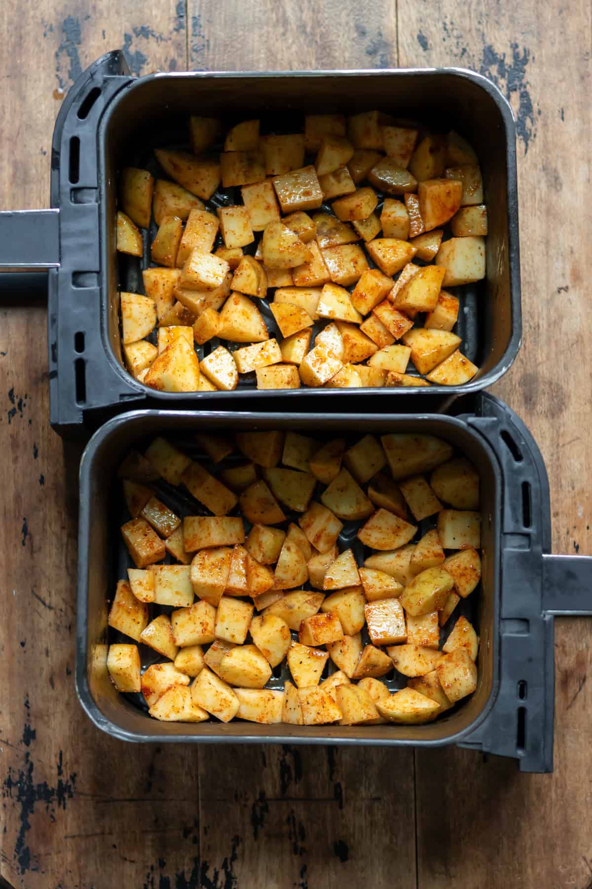 Seasoned diced potatoes in the baskets of an air fryer.