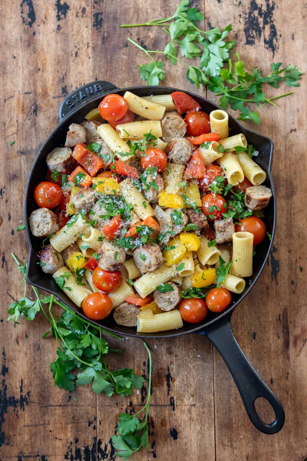 A wooden table with a cast iron skillet of Italian sausages and peppers, with parsley next to it.