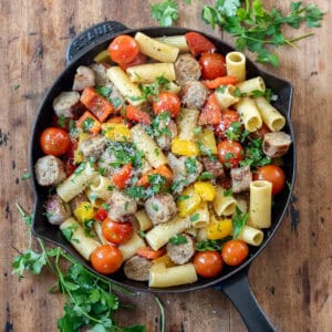 A wooden table with a cast iron skillet of Italian sausages and peppers, with parsley next to it.