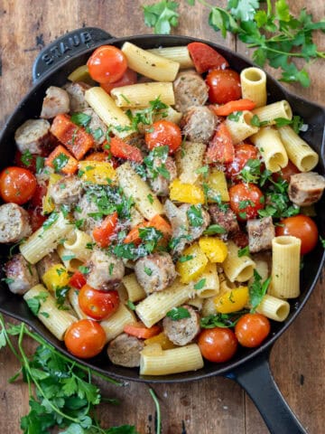 A wooden table with a cast iron skillet of Italian sausages and peppers, with parsley next to it.