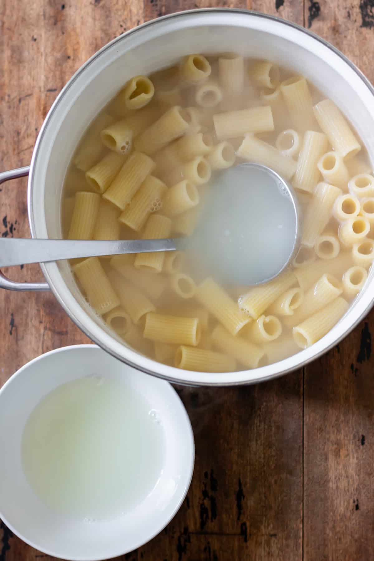 A pot of cooked rigatoni, with a ladle saving some of the cooking water in a bowl.