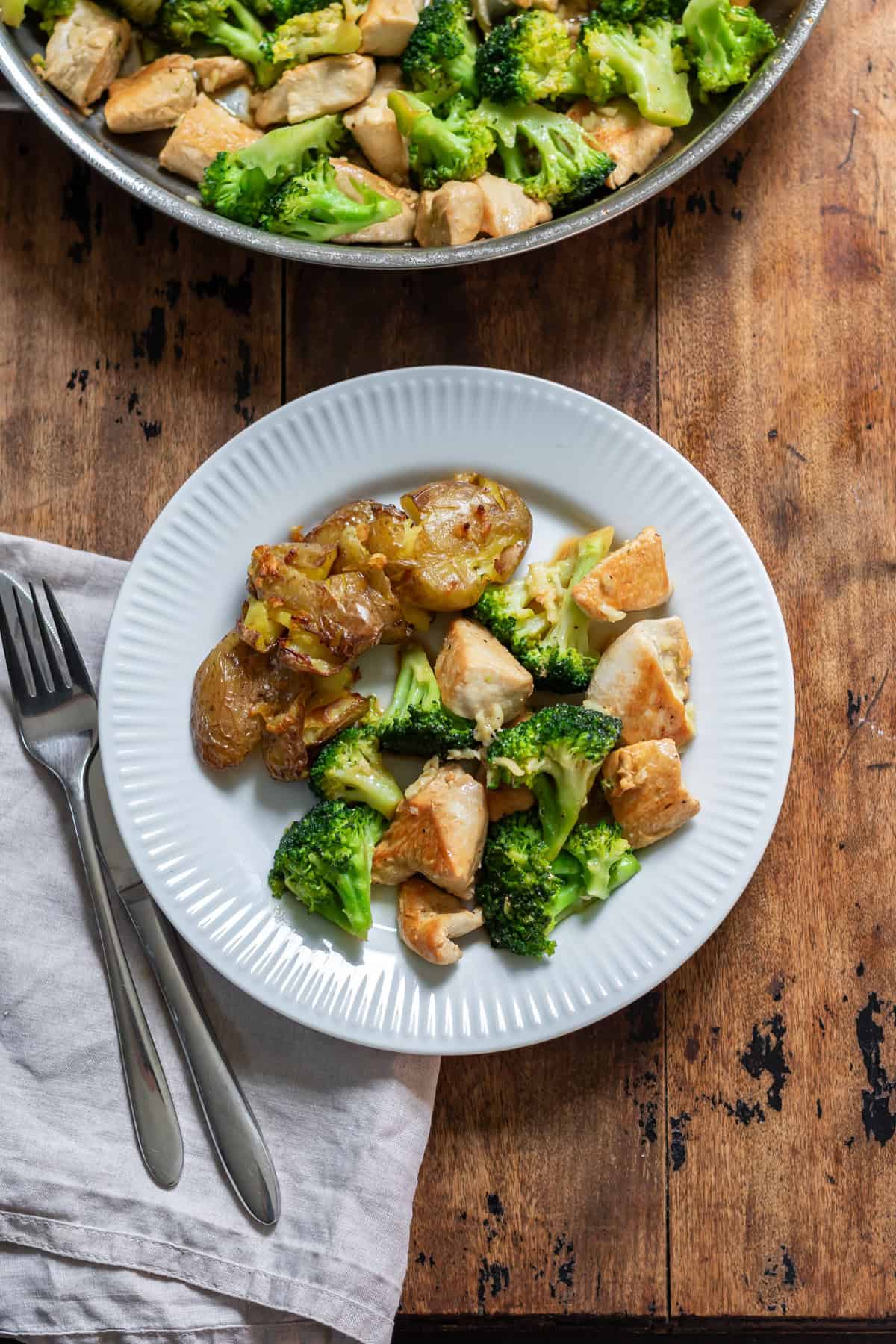 A wooden table with a plate of garlic butter chicken and broccoli skillet with roasted potatoes.