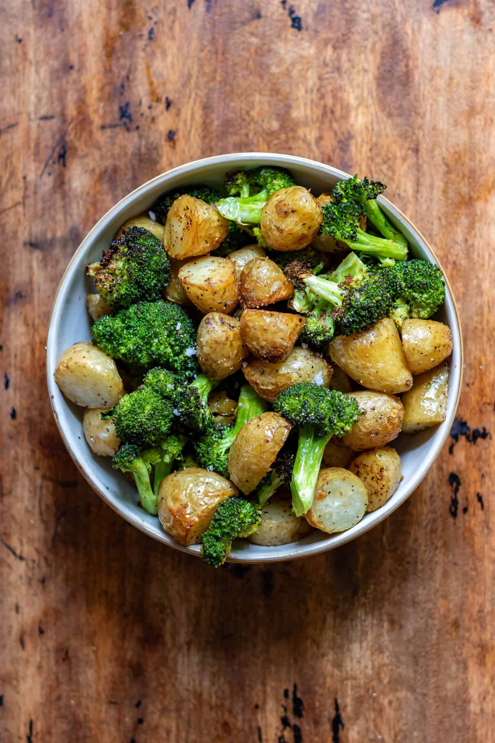 Wooden table with a bowl of garlic roasted broccoli and potatoes.