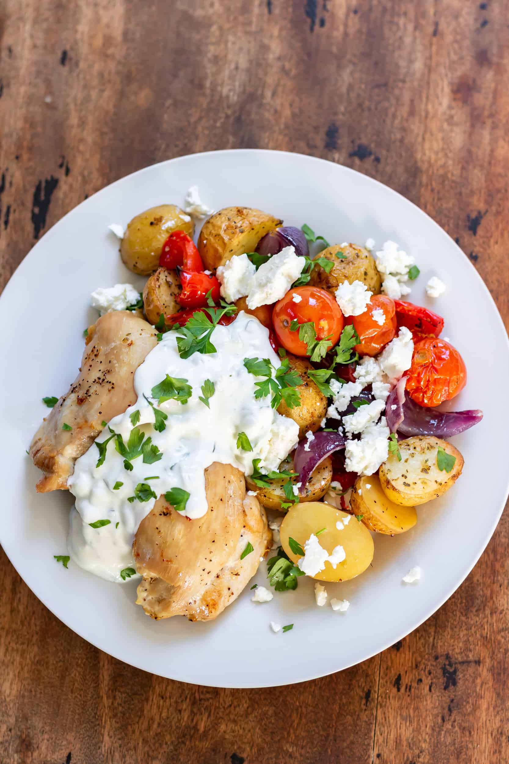 A plate with roasted Greek chicken, potatoes and vegetables, topped with tzatziki sauce.