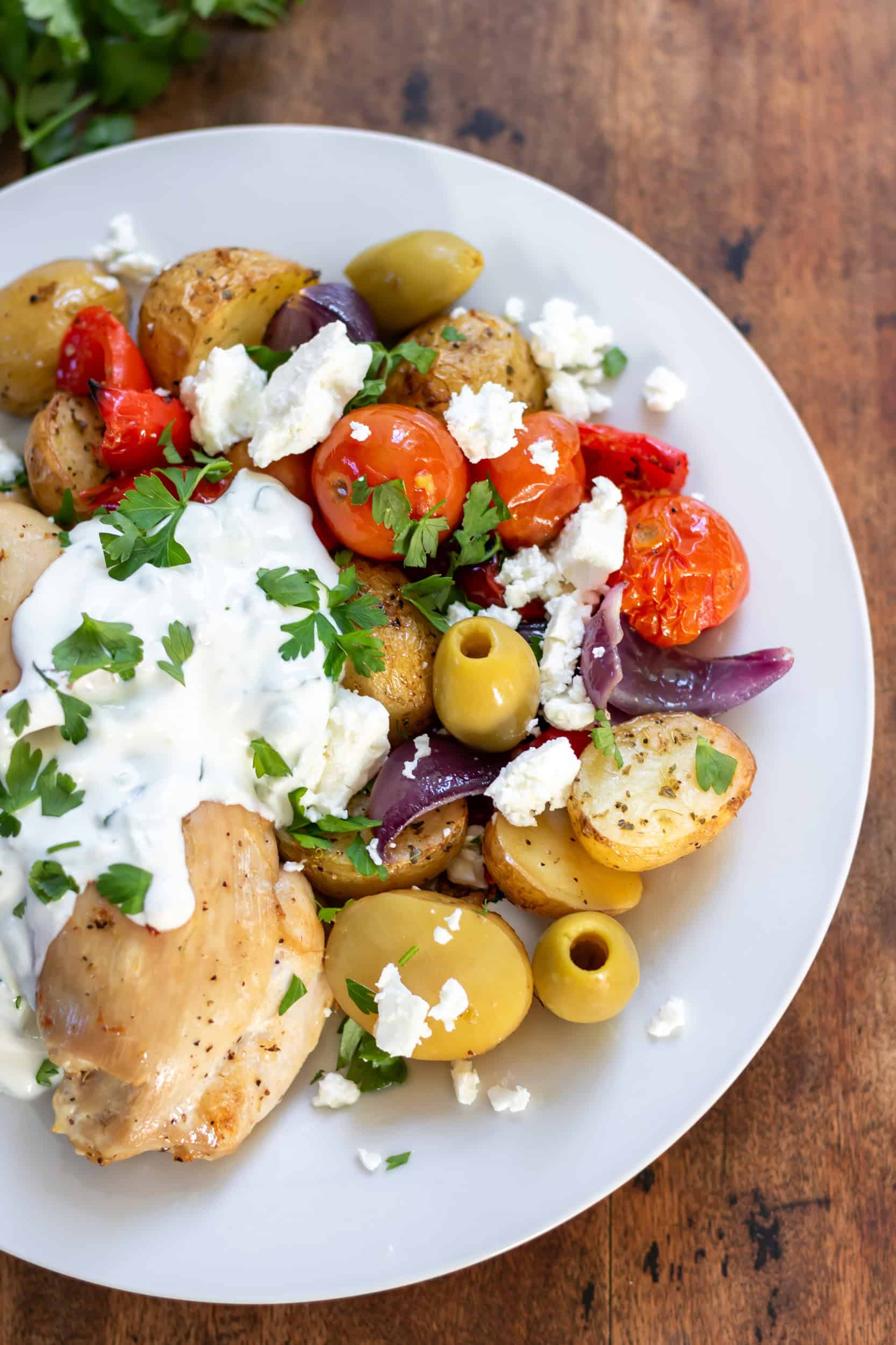 Close up of a plate with roasted Greek chicken thighs, potatoes, vegetables, topped with tzatziki sauce, feta, olives and parsley.