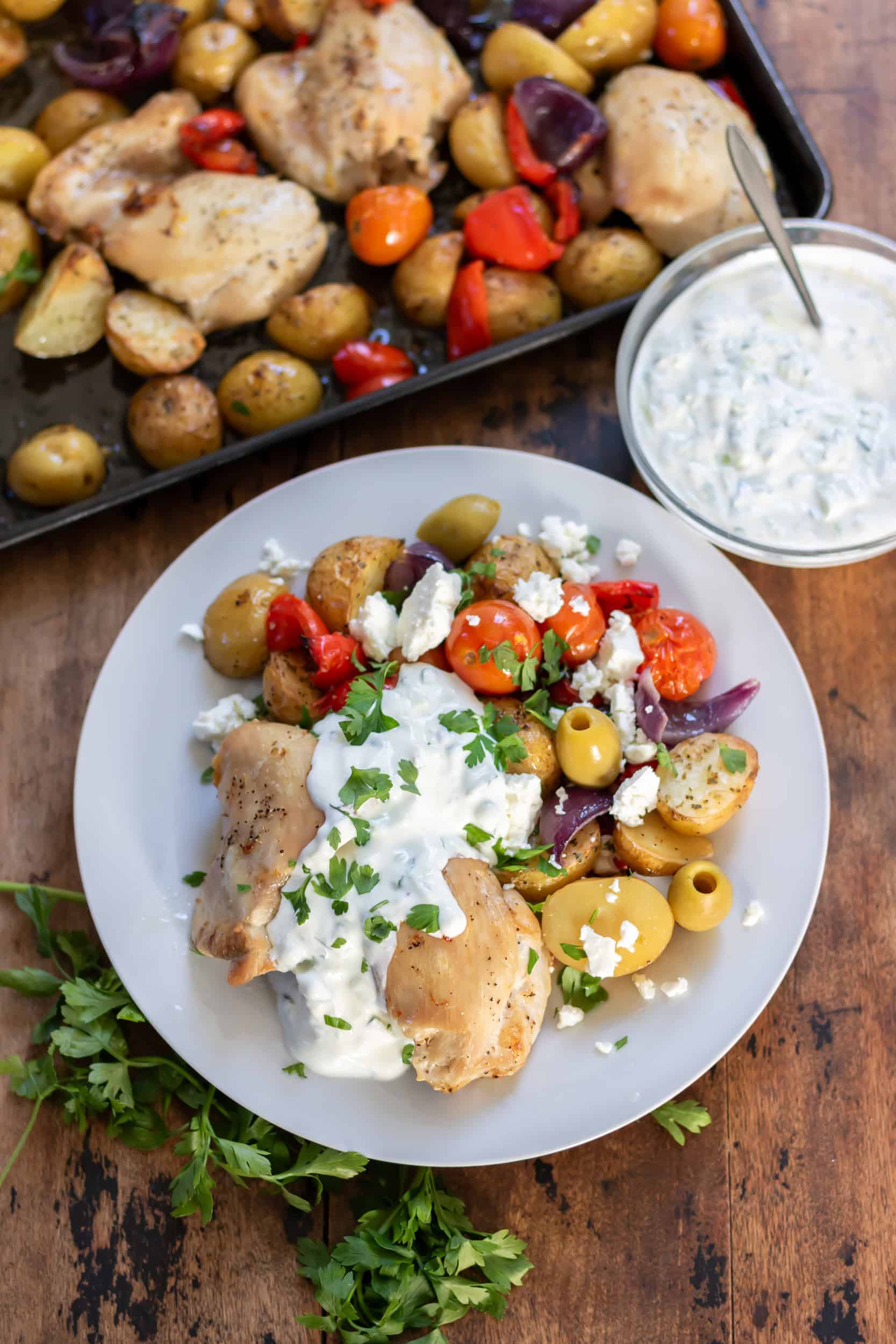 A table with a sheet pan of roasted chicken, potatoes and vegetables, with a portion on a plate.