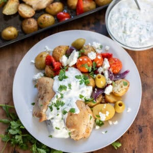 A plate with roasted Greek chicken thighs, potatoes, vegetables, topped with tzatziki sauce, feta, olives and parsley.