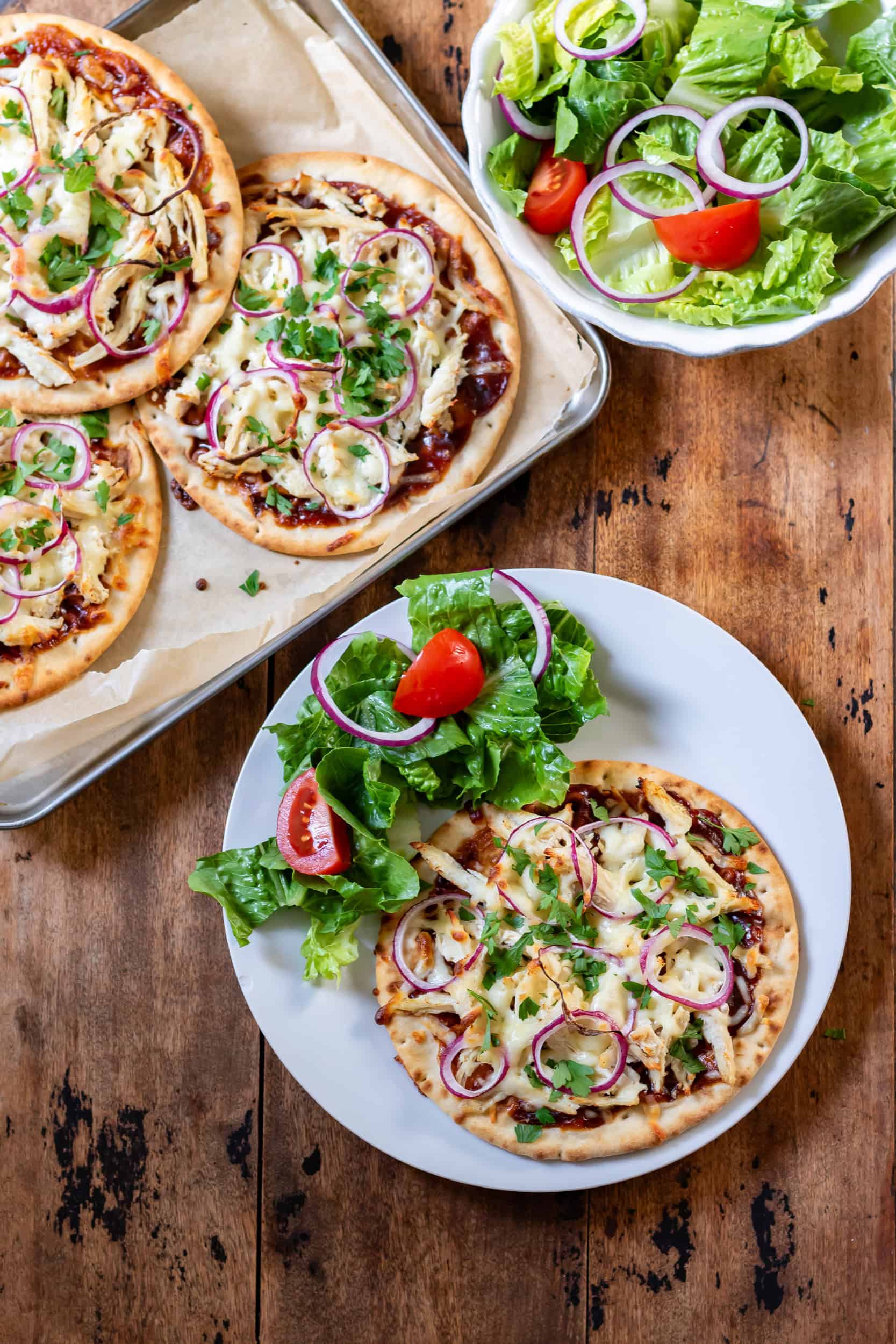 Looking down at a table with a baking sheet of bbq chicken flatbreads, a bowl of salad and a plate with a barbecue chicken flatbread and salad.