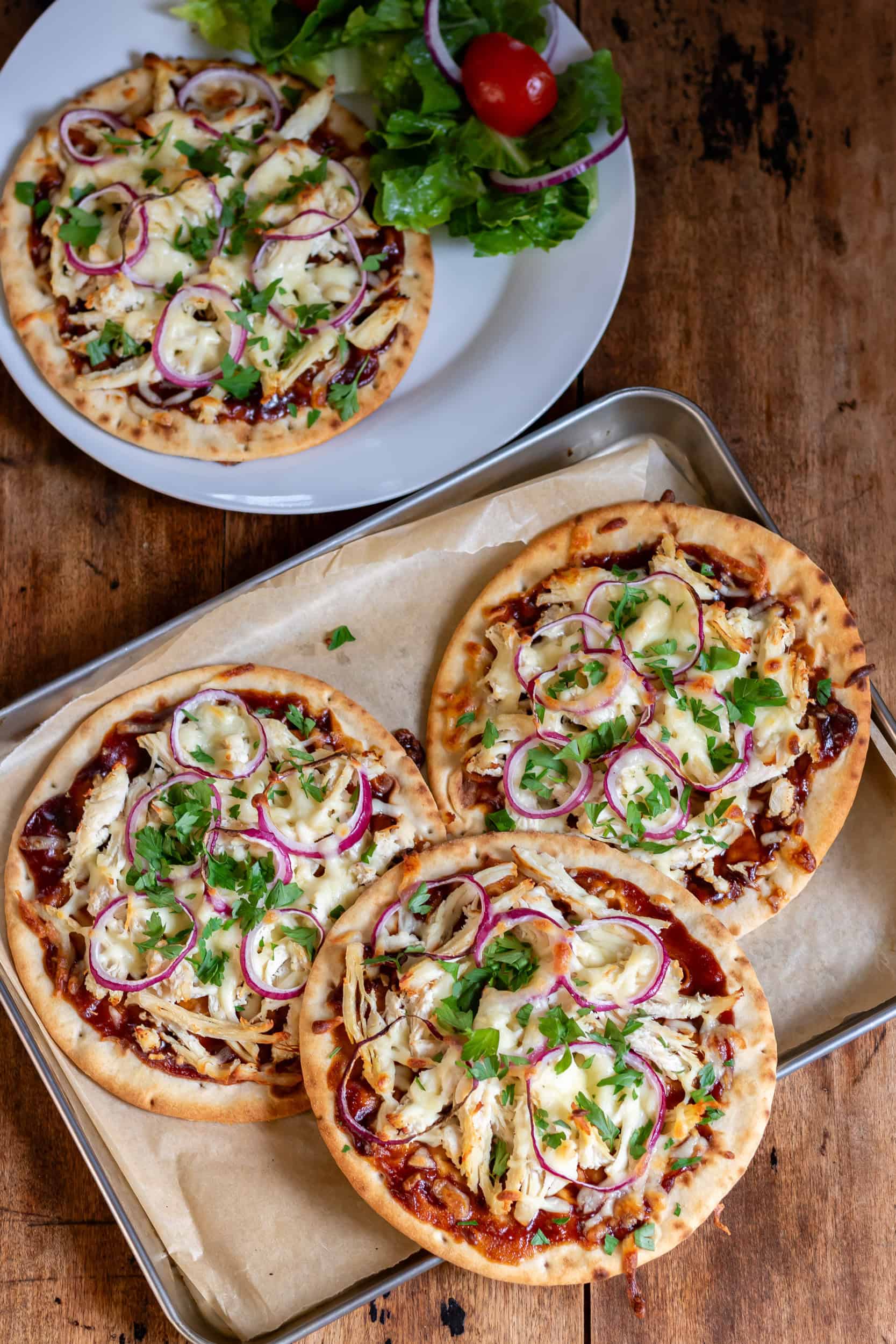 Looking down at a wooden table with a tray of three barbecue chicken flatbreads, plus a plate with a bbq chicken flatbread and salad.