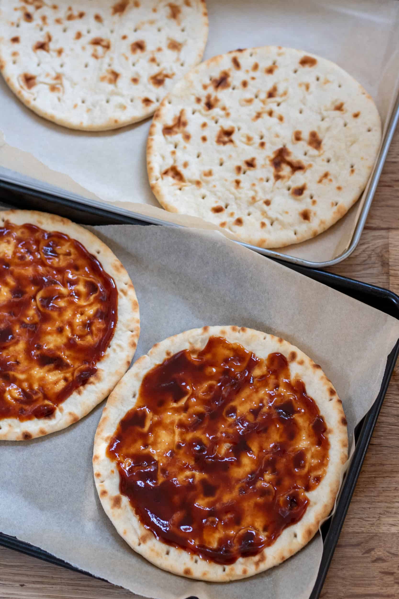Baking sheets with flatbreads being spread with barbecue sauce.