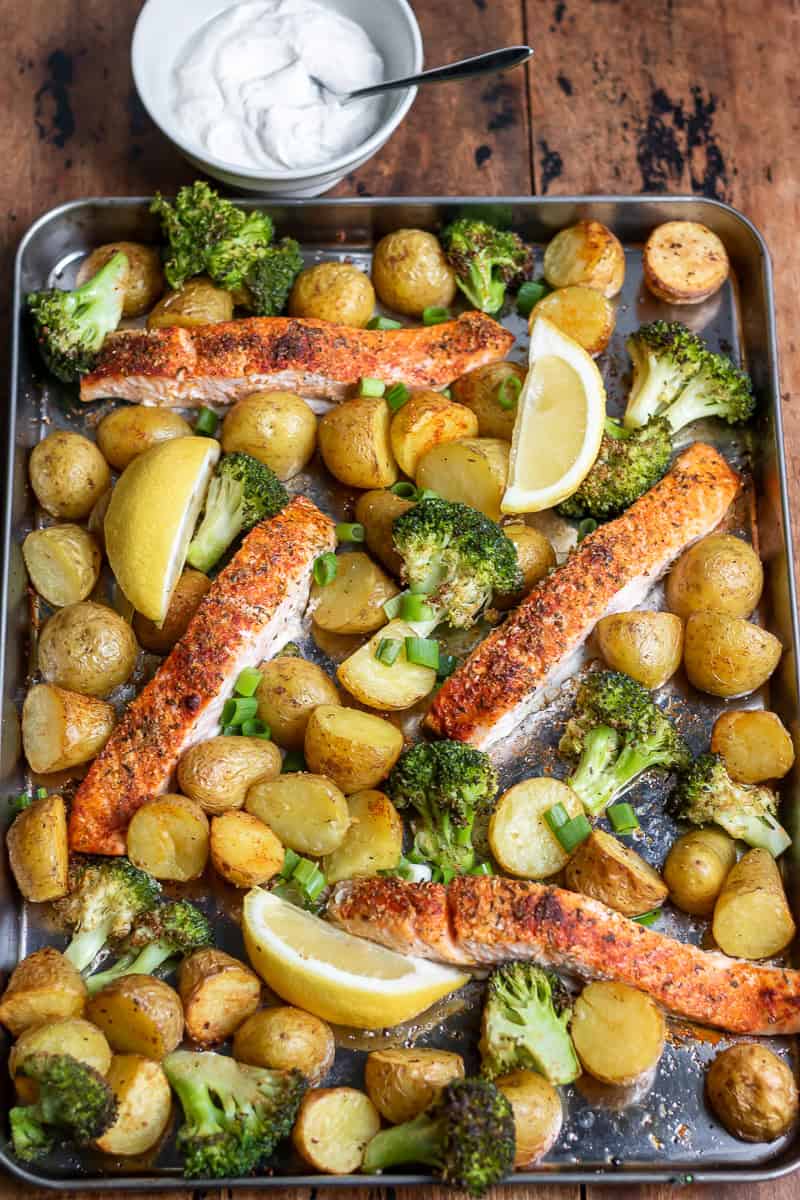 A table with a sheet pan salmon, broccoli and potatoes, next to a dish of horseradish yogurt sauce.