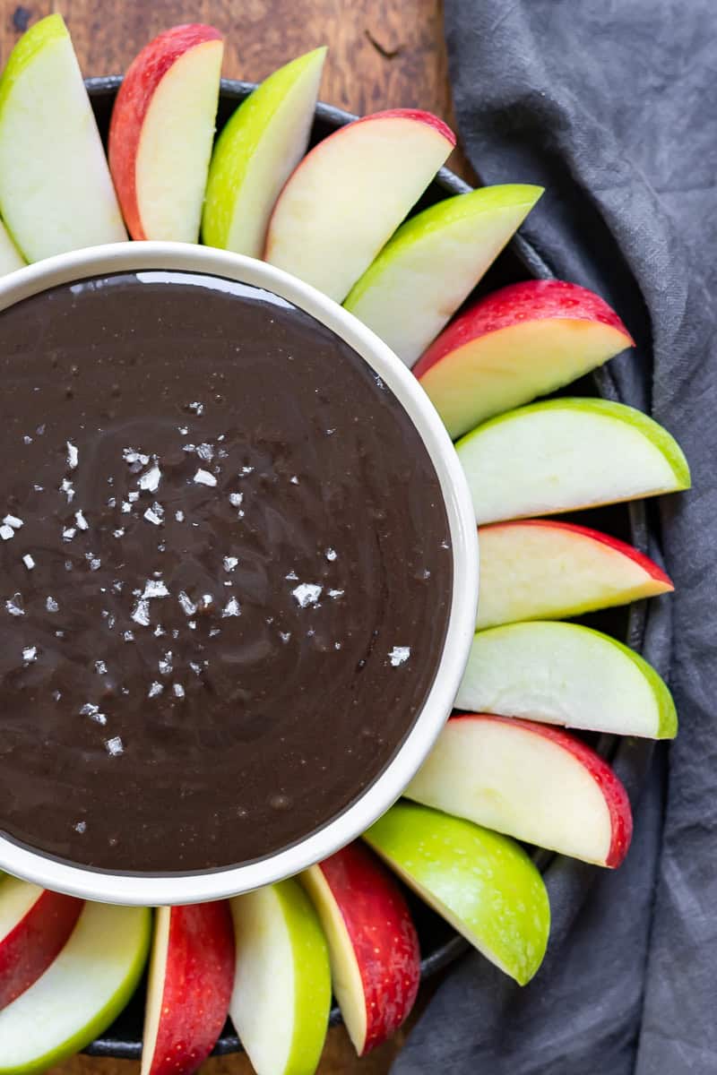 Close up of a bowl of black cocoa caramel dip with sea salt, surrounded by alternating sliced red and green apples.
