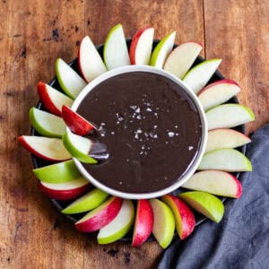 A wooden table with a bowl of halloween black cocoa caramel dip with apple slices dipped in.