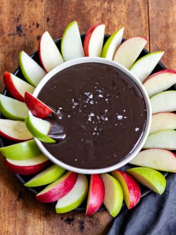 A wooden table with a bowl of halloween black cocoa caramel dip with apple slices dipped in.