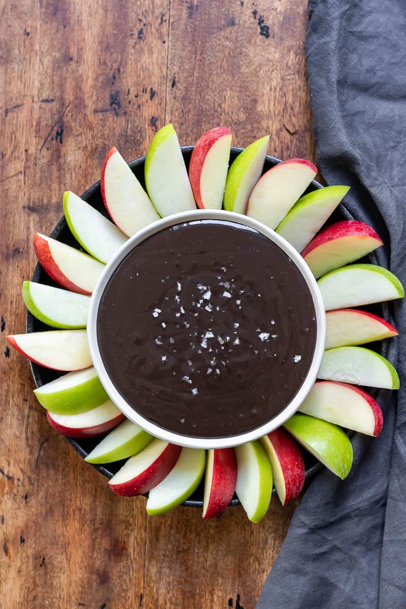 Bowl of black cocoa caramel dip with sea salt, surrounded by alternating sliced red and green apples.