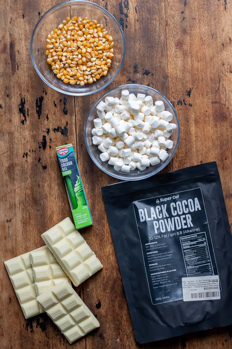 Popcorn, mini marshmallows, white chocolate, green food colouring and black cocoa on a table.